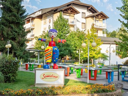 Familienhotel - Kinderhotels Europa - Maskottchen Sonnelino mit Hotel und Pit Pat im Hintergrund - Baby + Kinderhotel Sonnelino